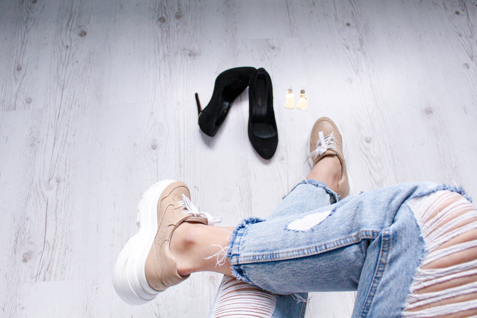 Tan sneakers and ripped jeans beside black high-heeled shoes and earrings on a pale wood floor.