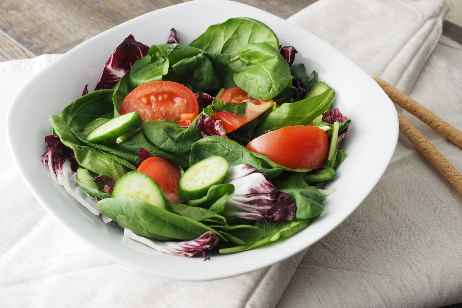 Leafy green salad with tomato and cucumber in a white bowl, with breadsticks beside it.