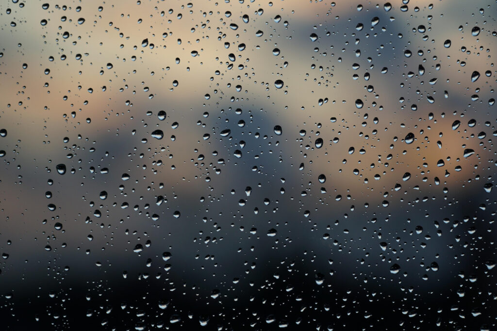 Raindrops on a window with a blurred outdoor view beyond the glass.